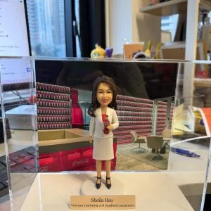 商務女性人像公仔身穿白色套裝內搭紅衣，手持玻璃瓶飲品，站在展示瓶罐牆與休憩區的企業背板前，前方有刻名銘牌並置於透明展示盒內