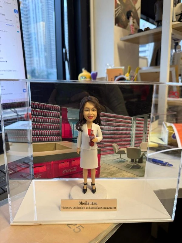 商務女性人像公仔身穿白色套裝內搭紅衣，手持玻璃瓶飲品，站在展示瓶罐牆與休憩區的企業背板前，前方有刻名銘牌並置於透明展示盒內