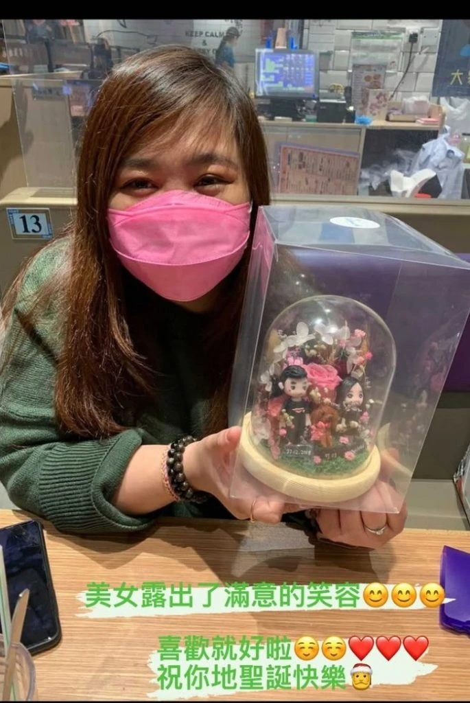 一位戴粉紅色口罩的女士，手持一個玻璃罩內的Q版真人公仔，公仔呈現一對情侶與一隻狗狗，周圍佈滿花卉，是讓客人露出滿意笑容的訂造人像公仔禮物。