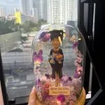 女生畢業公仔戴學士帽、手抱小玩偶與花束，立於紫粉永生花鋪陳的木座上，玻璃罩內點綴暖黃小燈並附有祝賀銘牌