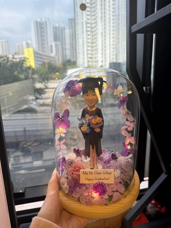 女生畢業公仔戴學士帽、手抱小玩偶與花束，立於紫粉永生花鋪陳的木座上，玻璃罩內點綴暖黃小燈並附有祝賀銘牌