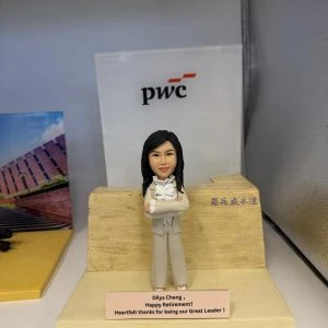 擺設上的女主管公仔:黑長髮、雙手交叉的女士穿米色套裝與荷葉邊襯衫,站在淺木石紋講壇前;前方銘牌寫有「Dilys Cheng, Happy Retirement! Heartfelt thanks for being our Great Leader!」,背景可見 pwc 標誌。