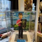 男士人像公仔穿紅色高爾夫上衣與黑長褲，手持「TOP 100 GOLF COURSE ASIA」獎牌站在六角磚地面上，背後是高爾夫球場風景背板，上有「The Jockey Club Kau Sai Chau Public Golf Course」字樣，前方藍色銘牌寫著 “Mike Carey Happy Retirement 2025”