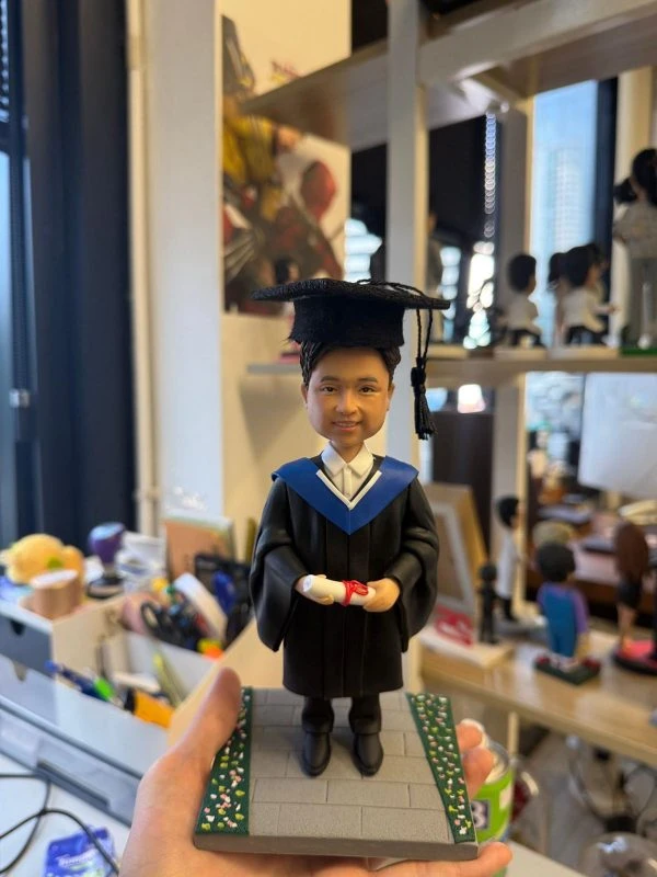 畢業主題人像公仔身穿黑色學袍與藍領披肩，頭戴學士帽，雙手捧著綁紅絲帶的畢業證書，站在兩側有小花點綴的灰色磚道底座上