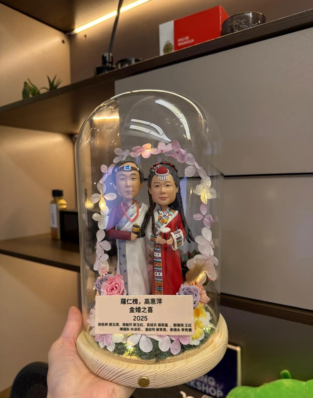 一對穿民族傳統禮服的夫妻人像公仔被裝在玻璃罩內，周圍環繞花環燈飾與木紋底座刻名牌