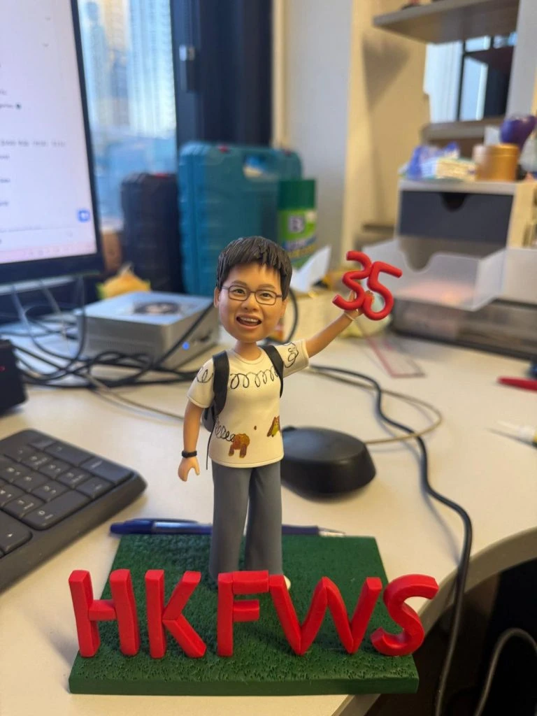 戴眼鏡的人像公仔背著背包、穿白色塗鴉T與灰色長褲，右手高舉紅色「35」，站在綠色草地底座，前方立體紅字「HKFWS」