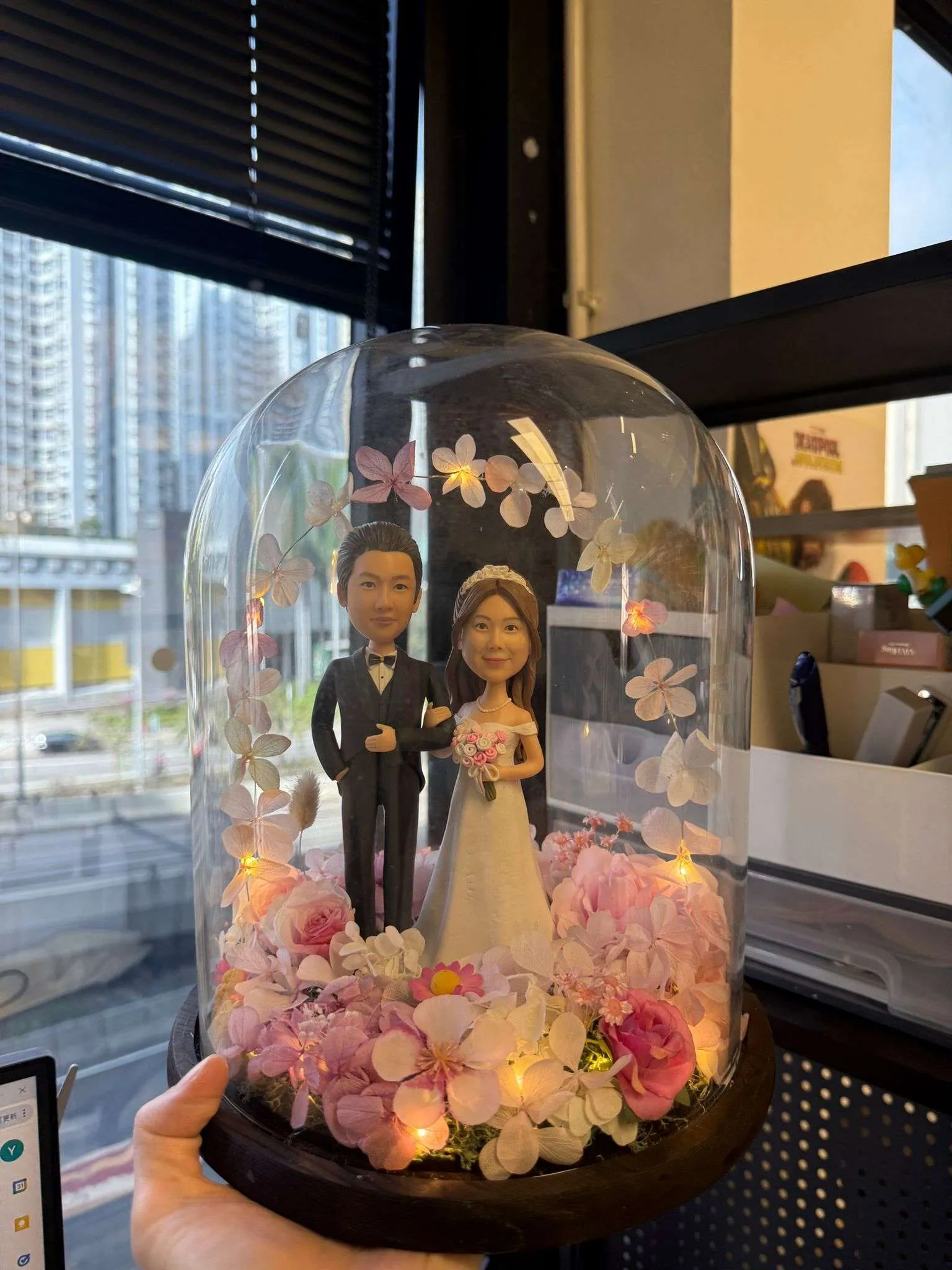 一對穿著婚紗禮服的訂造人像公仔，置於浪漫的粉色花卉玻璃罩內，配有小燈點綴，將新婚里程碑封存為永恆場景。