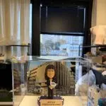 透明展示盒內的客製牧師公仔，女士手捧地球儀站立於紫色底座前，背板為教會建築外觀，前方銘牌刻有祝福與日期