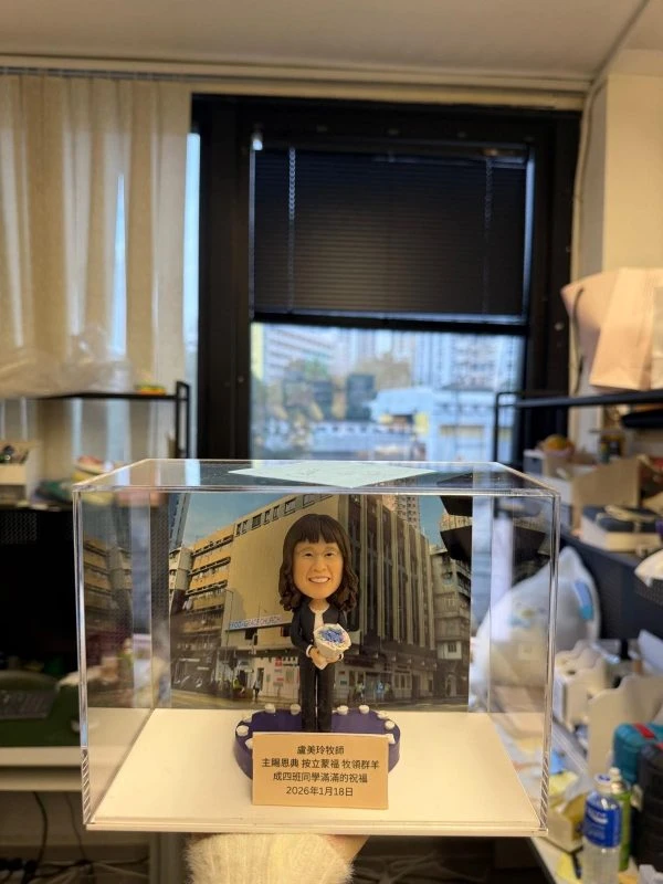 透明展示盒內的客製牧師公仔，女士手捧地球儀站立於紫色底座前，背板為教會建築外觀，前方銘牌刻有祝福與日期