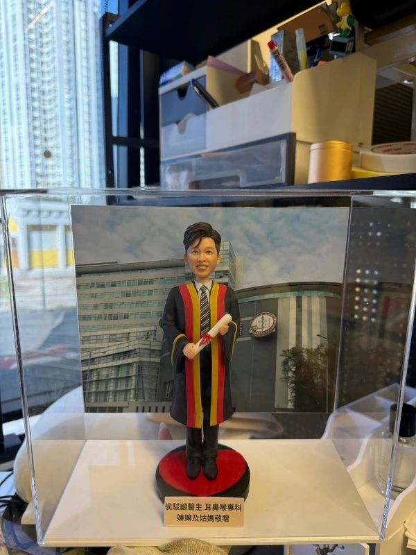 透明展示盒內的客製畢業公仔，男士穿黑色長袍配紅黃滾邊與領帶，手持捲軸站在紅黑底座上，背板為醫院大樓外觀，前方銘牌寫上致贈者訊息