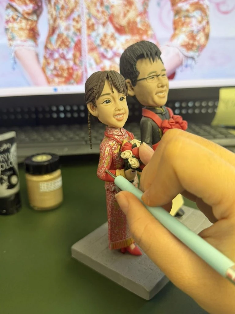 訂造人像公仔新娘穿著紅色龍鳳褂，手持花束，創作者正在為公仔的嫁衣細節進行刻畫，展現匠心獨運的工藝。