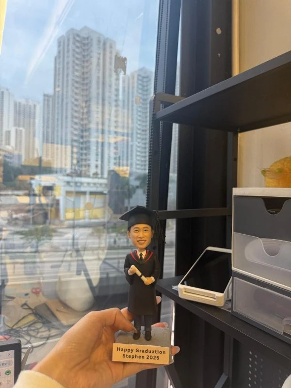 手持的客製畢業公仔，男生穿黑色學士袍與方帽、披紅邊披肩並拿著捲軸，底座銘牌寫「Happy Graduation Stephen 2025」