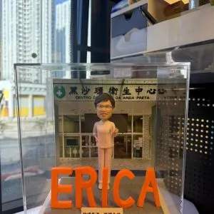 透明展示盒內的客製護士退休公仔，身穿粉色護理服站在醫療中心門前，前方以大型橙色字母擺出「ERICA」，底座銘牌刻有祝福語與贈送者姓名