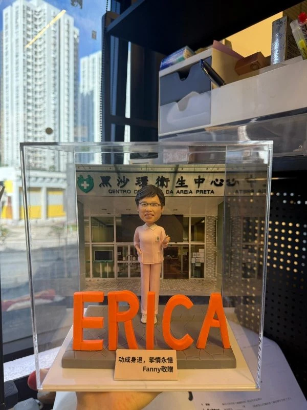 透明展示盒內的客製護士退休公仔，身穿粉色護理服站在醫療中心門前，前方以大型橙色字母擺出「ERICA」，底座銘牌刻有祝福語與贈送者姓名