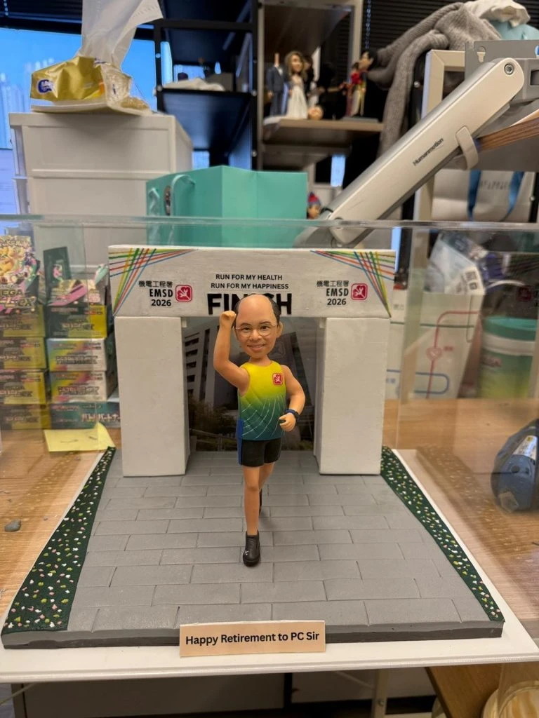 一個戴眼鏡嘅Q版真人公仔穿著彩色運動背心和黑色短褲，站在馬拉松終點線前，場景牌寫上「Happy Retirement to PC Sir」，是送俾上水警署師兄嘅榮休人像公仔。