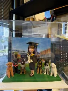 透明展示盒中的客製畢業公仔，戴方帽穿黑色學袍的女生抱著一隻小狗，左右排站多隻不同品種的狗，背板為校園建築