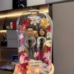 一對 Q版真人公仔，穿著日常服裝，分別抱著黑色貴婦狗，在玻璃罩內以永生花和燈飾佈置，祝賀長輩光榮退休。