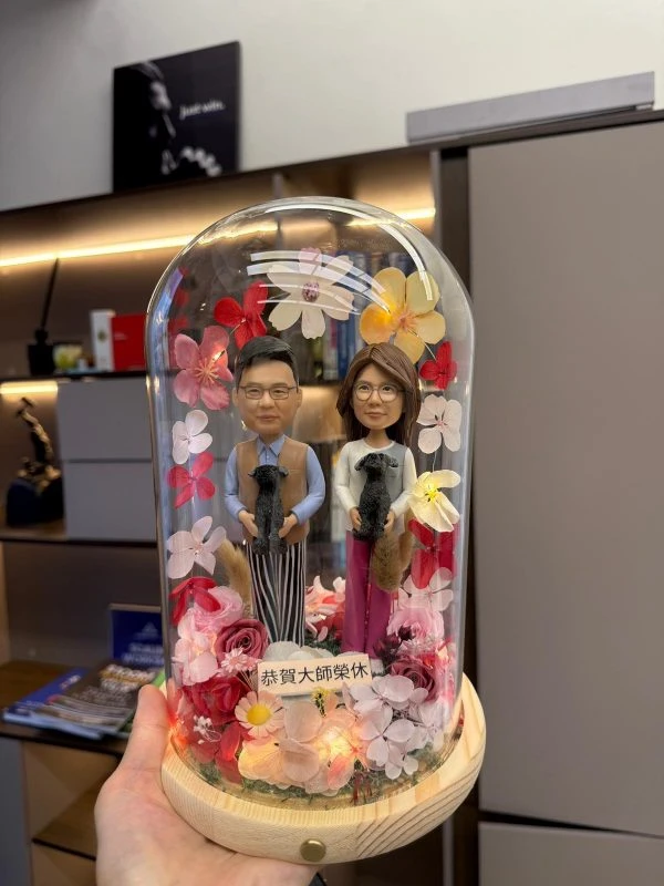 一對 Q版真人公仔，穿著日常服裝，分別抱著黑色貴婦狗，在玻璃罩內以永生花和燈飾佈置，祝賀長輩光榮退休。