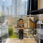透明展示盒內的客製醫護公仔，穿深藍手術服雙手交叉站立，背板為現代醫學院建築，底座銘牌寫有「Dr. Veronica Lam School of Nursing, HKU」