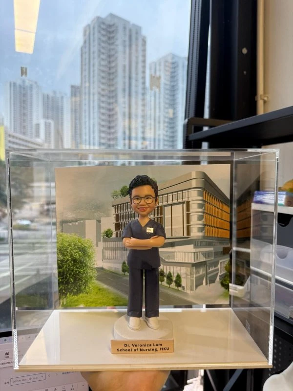 透明展示盒內的客製醫護公仔，穿深藍手術服雙手交叉站立，背板為現代醫學院建築，底座銘牌寫有「Dr. Veronica Lam School of Nursing, HKU」