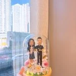 玻璃花盅內的一對客製情侶公仔，女方穿深色花裙抱白色小狗，男方穿黑色襯衫抱黑色小狗，周圍以粉玫瑰、白花與暖光燈點綴，木質底座襯草地