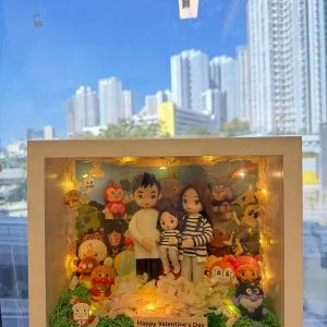 手持白色立體相框式展示盒，內有一家三口公仔：大人兩位與小孩一位，穿著黑白條紋與淺色服裝，前方鋪綠草與淺色花材；四周與背板佈滿多個可愛卡通角色與小擺件，盒內繞有暖黃燈串；前方小牌寫著「Happy Valentine’s Day」。背景是窗外藍天與高樓。