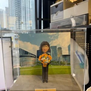 透明展示盒中的客製公仔，身穿黑色長褲與橘色元素的上衣，雙手捧著卡通造型花束；背板是草地與現代建築群的風景圖；前方金色小名牌寫有「Apple Happy Retirement 2026」；擺放在室內桌面前，背景可見辦公室收納物與窗外高樓。