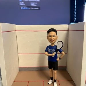 玻璃纖維或黏土製的客製公仔站在迷你壁球場景中，穿藍色運動上衣與黑色短褲，手持球拍，腳邊有一顆黑色壁球，背景是三面白牆與紅色界線；遠處可見電腦螢幕的模糊畫面。