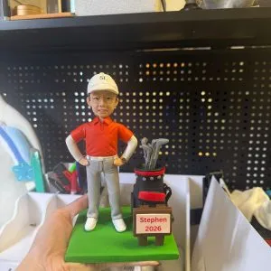 一個以高爾夫為主題的客製化人像公仔,人物神態自然,搭配球袋與草地底座,適合作為退休或紀念收藏。