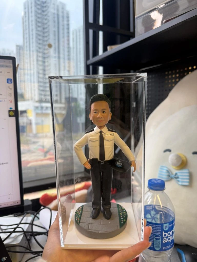 手持透明展示盒的近景照：盒內是一位身穿淺色制服襯衫、黑領帶與黑長褲的警務主題公仔，右手叉腰、左手持警帽；腳下為圓形灰色地磚基座，邊緣點綴綠地與小花紋；背景為辦公室桌面與窗外高樓，右側可見一瓶礦泉水。