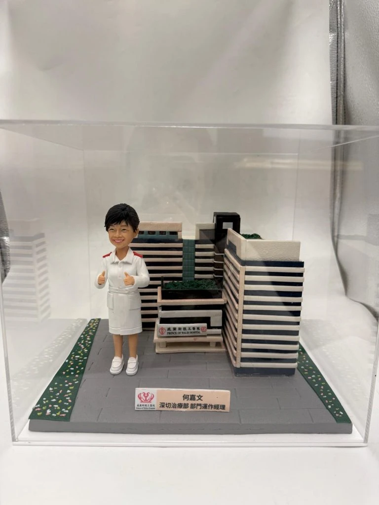 透明展示盒內的醫療主題客製公仔，身穿白色制服與白鞋、雙手比讚站在灰色地台上；旁邊有縮小版建築與花園模型，前方小牌寫有中文姓名與「深切治療部門運作經理」等字樣並帶有紅色徽標；地台兩側有綠色草地與小花紋路；背景為白色布幕。