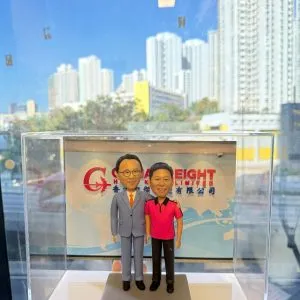 透明展示盒中的雙人客製公仔並肩站立，左側穿淺灰西裝配橘色領帶，右側穿桃紅與黑色拼接的休閒上衣與黑長褲；腳下為灰色長方形基座；背板印有公司英文縮寫與中文公司名稱的圖樣；背景透過玻璃可見高樓林立的城市景色與藍天。