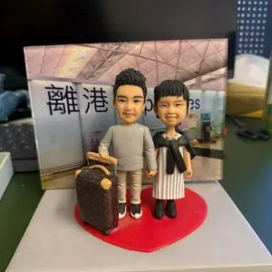 CUTEFIGUREHK 客製化情侶人像公仔，男女主角站在紅色心形底座上，男主角手扶精緻行李箱，配搭香港國際機場「離港 Departures」背景板，是專為親友送行訂製的移民禮物與送別禮物。