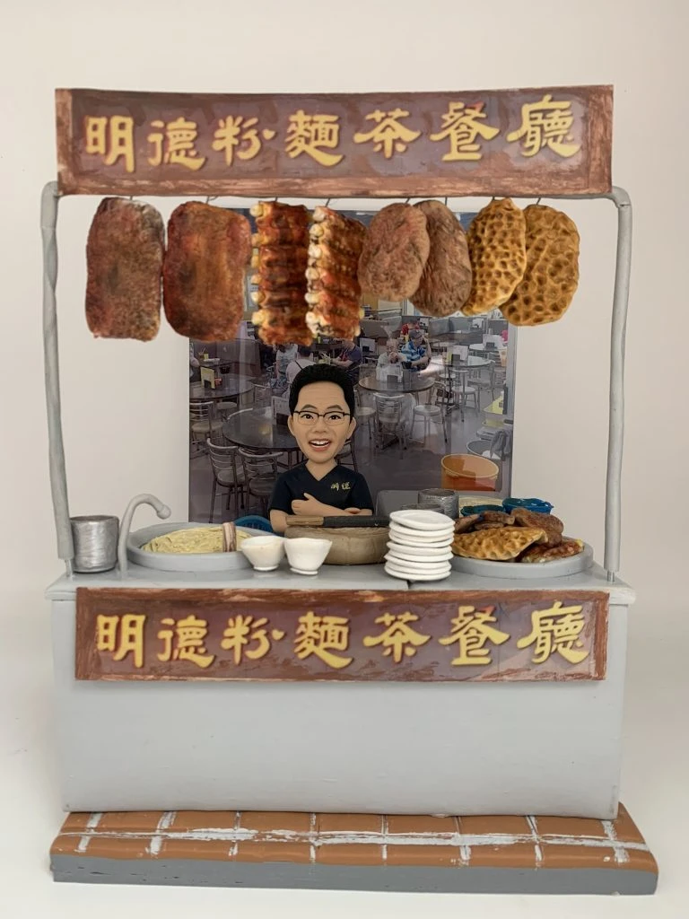 一個Q版真人公仔站在迷你茶餐廳攤位前，攤位掛著「明遠粉麵茶餐廳」招牌，並掛有燒味和格仔餅，象徵Modi Hair的創業夢想。
