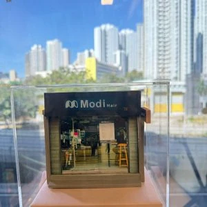 CUTEFIGUREHK 客製化店舖微縮模型。一個精緻的微型髮型屋店面模型,頂部有黑底藍字及白字的招牌寫著「Modi Hair」和「安寧路49號」。店面採用透明玻璃門設計,內部貼有價目表海報,隱約可見店內的裝潢和一張黃色高腳椅。模型放置在一個淺粉紅色的方形底座上,並罩著透明的壓克力展示盒。背景是隔著玻璃窗看到的城市高樓大廈和藍天。