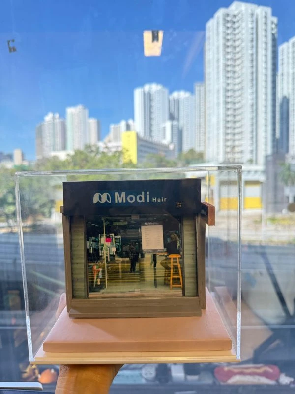 CUTEFIGUREHK 客製化店舖微縮模型。一個精緻的微型髮型屋店面模型，頂部有黑底藍字及白字的招牌寫著「Modi Hair」和「安寧路49號」。店面採用透明玻璃門設計，內部貼有價目表海報，隱約可見店內的裝潢和一張黃色高腳椅。模型放置在一個淺粉紅色的方形底座上，並罩著透明的壓克力展示盒。背景是隔著玻璃窗看到的城市高樓大廈和藍天。