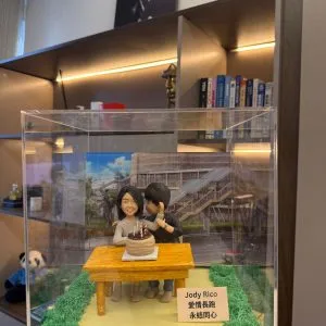 CUTEFIGUREHK 客製化情侶場景公仔。壓克力展示盒內，一對情侶坐在戶外木桌前慶祝生日，桌上有插著蠟燭的蛋糕。男主角親暱靠近女主角，背景為城市街道照片。底座有草皮裝飾及寫著「Jody Rico 愛情長跑 永結同心」的名牌，是完美的週年或情人節紀念禮物。