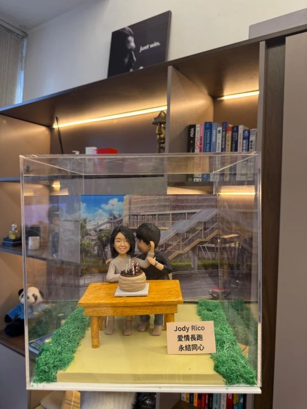CUTEFIGUREHK 客製化情侶場景公仔。壓克力展示盒內，一對情侶坐在戶外木桌前慶祝生日，桌上有插著蠟燭的蛋糕。男主角親暱靠近女主角，背景為城市街道照片。底座有草皮裝飾及寫著「Jody Rico 愛情長跑 永結同心」的名牌，是完美的週年或情人節紀念禮物。