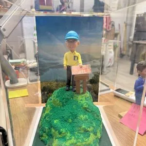 CUTEFIGUREHK 客製化行山主題紀念公仔。一位戴著淺藍色鴨舌帽和眼鏡的男士，身穿胸前印有藍色英文字母「D」的黃色Polo恤和深色長褲，面帶笑容地站在一個立體的綠色草地山頂模型上。他身旁立著一塊寫有「仙姑峰」（Hsien Ku Fung）及海拔高度的仿真木牌。公仔背後是一幅壯麗的香港山水風景（八仙嶺及船灣淡水湖）背景圖。整個模型被精心放置在一個透明的亞加力膠展示盒中。