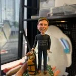 CUTEFIGUREHK 客製化男士人像公仔，生動還原了身穿深色 L.L.Bean 上衣及牛仔褲的男士，手拖著放有旅行袋的黃色行李箱。公仔站立於帶有草地小徑的底座上，前方設有寫著「Heartfelt thanks for being our great leader」的專屬感謝牌。這是一份極具心思、專為感謝上司或領導而設的散水、升職或榮休紀念禮物。