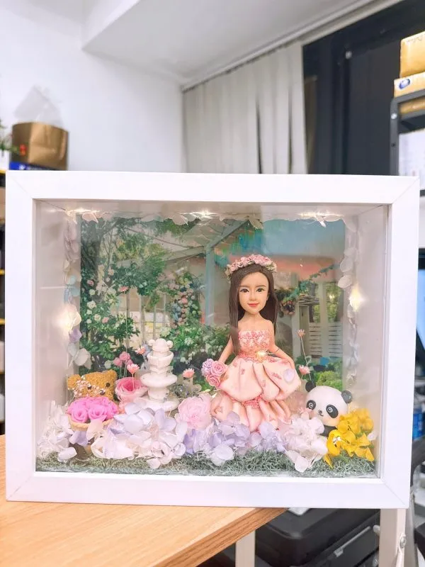 CUTEFIGUREHK 客製化立體相框公仔禮物。一個白色的發光畫框，內部佈置成夢幻的童話花園。中央是一個穿著粉紅色花瓣晚裝的長髮女孩立體公仔，頭戴花環，手持粉紅玫瑰。背景是浪漫的黃昏花園圖案，公仔腳下鋪滿了紫色、白色和粉紅色的乾花（繡球花）以及綠色苔蘚，旁邊點綴著微縮的白色小噴泉、可愛的小棕熊和熊貓模型。相框內置有溫馨的 LED 燈串，營造出唯美的氣氛。