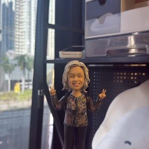 CUTEFIGUREHK 客製化女性人像公仔,生動還原了人物的灰髮、眼鏡造型以及雙手舉V字的開心神態,身穿花紋上衣並揹著背囊。公仔底座設有專屬牌匾,刻上「鄭姑娘 一無掛慮 平安喜樂❤️2025」的溫馨祝福語,是一份為同事度身訂造的退休或散水紀念禮物。