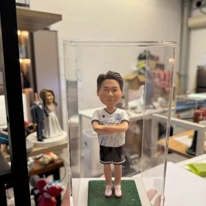 一尊裝在透明壓克力展示盒內的客製化男性足球員立體公仔。公仔雙手交叉於胸前,身穿印有「GIN BOUTIQUE」字樣與隊徽的白色足球球衣、黑色短褲(褲管印有數字14)以及紅白相間的球鞋。公仔站在綠色仿草皮質感的底座上,展現出運動員的自信風采。背景為擺放著其他公仔作品(如婚紗公仔)的工作室環境。