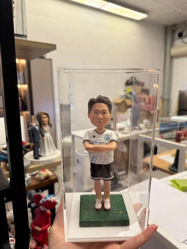 一尊裝在透明壓克力展示盒內的客製化男性足球員立體公仔。公仔雙手交叉於胸前，身穿印有「GIN BOUTIQUE」字樣與隊徽的白色足球球衣、黑色短褲（褲管印有數字14）以及紅白相間的球鞋。公仔站在綠色仿草皮質感的底座上，展現出運動員的自信風采。背景為擺放著其他公仔作品（如婚紗公仔）的工作室環境。