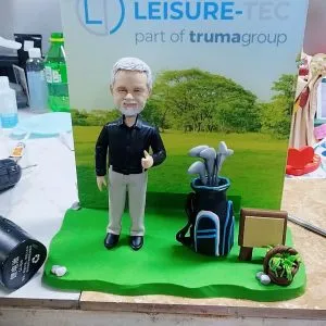 CUTEFIGUREHK 客製化外籍上司榮休公仔。白髮白鬚的男士身穿黑色襯衫與淺色長褲，比出讚好手勢，身旁配有精緻的藍黑色高爾夫球袋及球桿微縮模型。背板印有「LEISURE-TEC」公司標誌及戶外大自然風景，綠色草地底座上設有小木牌及花籃點綴，是送給熱愛高爾夫球的老闆或高管的最佳退休紀念禮物。