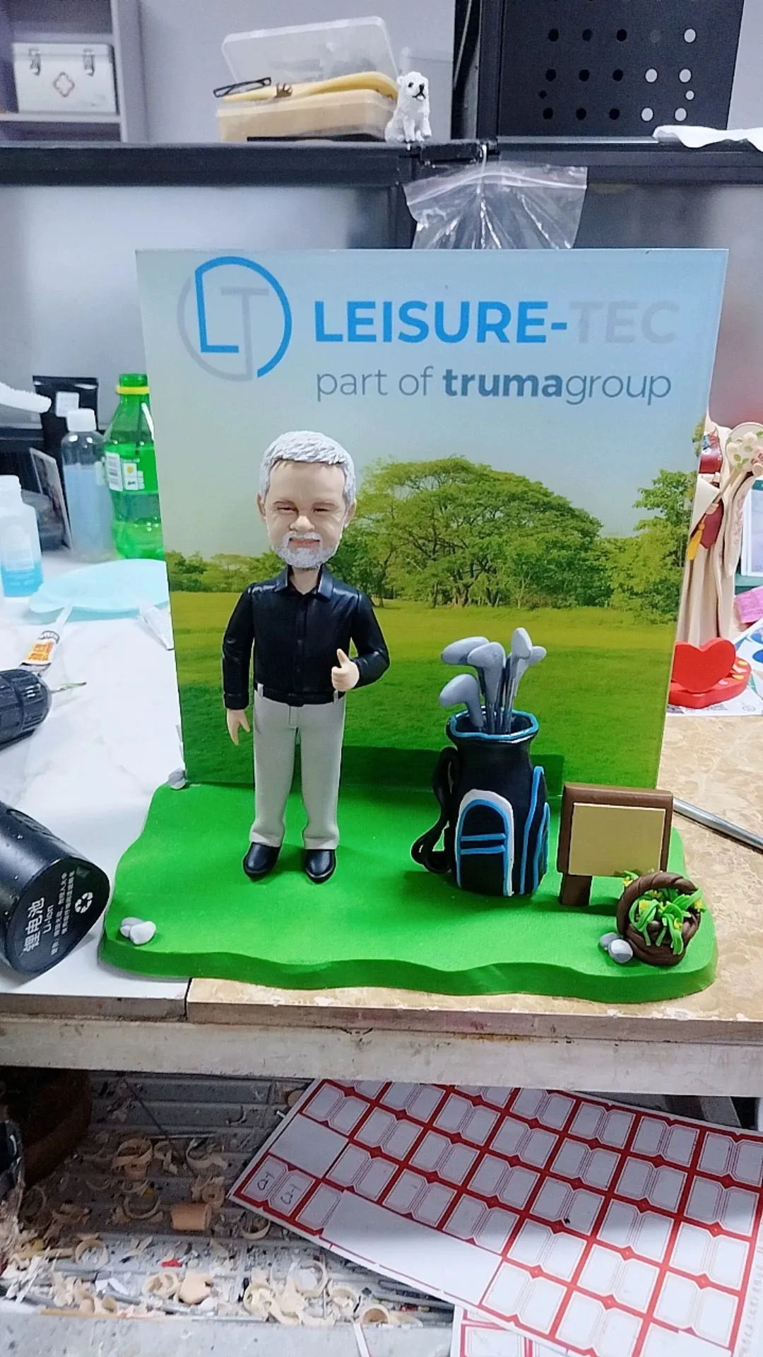 CUTEFIGUREHK 客製化外籍上司榮休公仔。白髮白鬚的男士身穿黑色襯衫與淺色長褲，比出讚好手勢，身旁配有精緻的藍黑色高爾夫球袋及球桿微縮模型。背板印有「LEISURE-TEC」公司標誌及戶外大自然風景，綠色草地底座上設有小木牌及花籃點綴，是送給熱愛高爾夫球的老闆或高管的最佳退休紀念禮物。