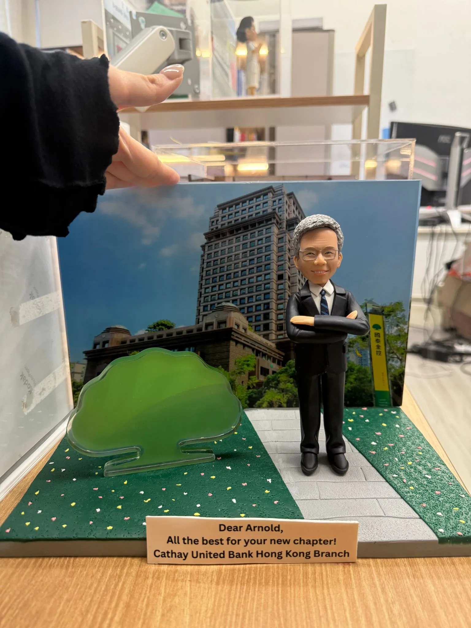 CUTEFIGUREHK 客製化企業紀念公仔。公仔為一位銀髮、戴眼鏡、身穿深色西裝打領帶的男士，雙手交叉抱胸，展現專業自信。場景配有國泰金控大樓背景圖及標誌性的綠色大樹道具。底座名牌刻有「Dear Arnold, All the best for your new chapter! Cathay United Bank Hong Kong Branch」，是一份專為銀行高管訂製的榮休或散水禮物。