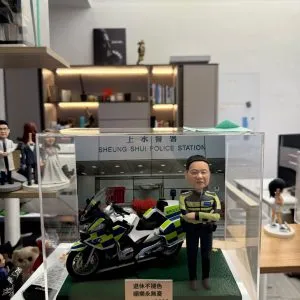CUTEFIGUREHK 客製化香港交通警公仔。公仔身穿螢光黃綠色交通警制服，雙手交叉抱胸。旁邊配有一架精緻的警察電單車模型。場景背景為「上水警署」(Sheung Shui Police Station)。底座名牌刻有「退休不褪色 順樂永無憂 2026」，是一份專為警察訂製的榮休紀念禮物，裝在透明亞加力展示盒中。