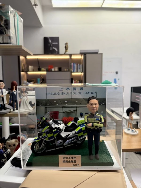 CUTEFIGUREHK 客製化香港交通警公仔。公仔身穿螢光黃綠色交通警制服，雙手交叉抱胸。旁邊配有一架精緻的警察電單車模型。場景背景為「上水警署」(Sheung Shui Police Station)。底座名牌刻有「退休不褪色 順樂永無憂 2026」，是一份專為警察訂製的榮休紀念禮物，裝在透明亞加力展示盒中。
