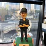 CUTEFIGUREHK 客製化香港海關人員人像公仔。公仔生動還原了人物的面貌，並穿著整齊的深藍色海關制服與海關帽，雙手交叉胸前，展現專業自信的姿態。公仔站立於點綴了小花的綠色底座上，前方設有印著「總關員 黃文傑 榮休之喜」的專屬紀念牌。這是一份專為紀律部隊人員度身訂造的退休致敬禮物。